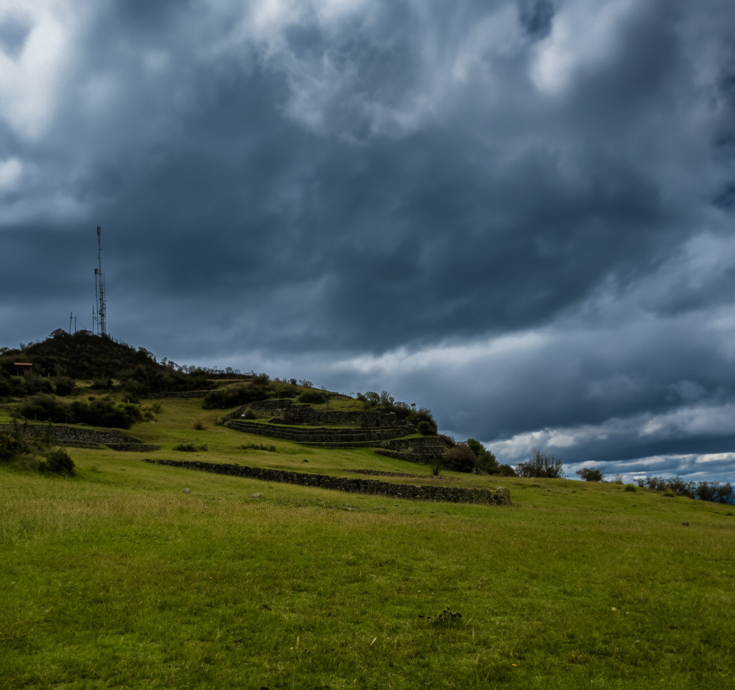 Edit this existing photo of an Ecuadorian Andean hillside with stone terraces and a communications tower. Two specific changes: 1) LANDSCAPE below the horizon: brighten and enhance the green hills, grass, and stone terrace walls — make them sharper, more vivid and clear, with better contrast and detail, as if shot with a high-resolution camera. More color saturation in the greens, clearer stone textures. 2) SKY above the horizon: keep a dramatic dark blue-grey stormy sky — heavy brooding storm clouds, dark slate blue-grey moody tones, completely overcast. Absolutely NO rays of light, NO sunbeams, NO golden glow, NO bright spots anywhere. The contrast between the bright clear landscape below and the dark dramatic sky above should feel striking.